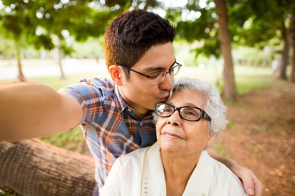 Grandmother seated in a park with young teen grandson next to her, kissing her forehead