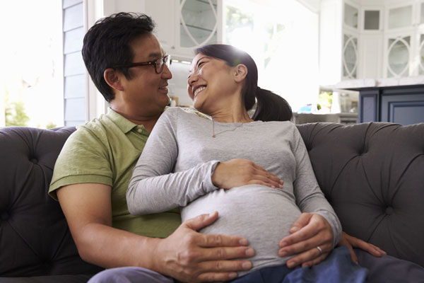 Happy young Asian couple seated on couch, smiling at each other, wife is pregnant