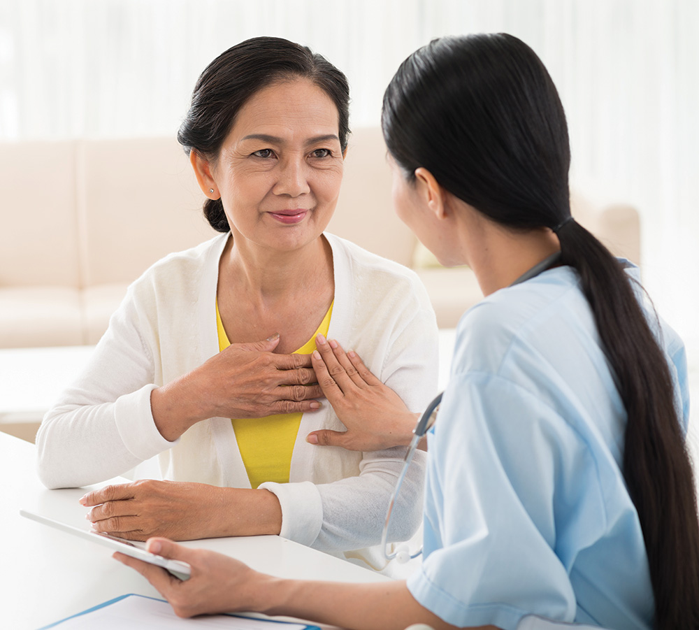 Seated nurse, with long dark ponytail, shows fully clothed seated middle age Asian woman how to do a breast self-exam