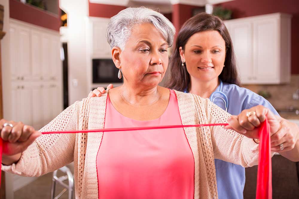 Older woman being shown by a young nurse how to do a stretch band exercise with her arms