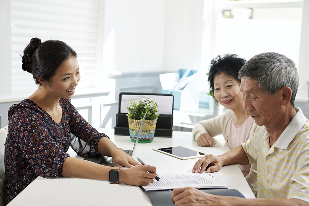 Smiling hospital relations woman speaking with middle aged Asian couple, all seated at a table