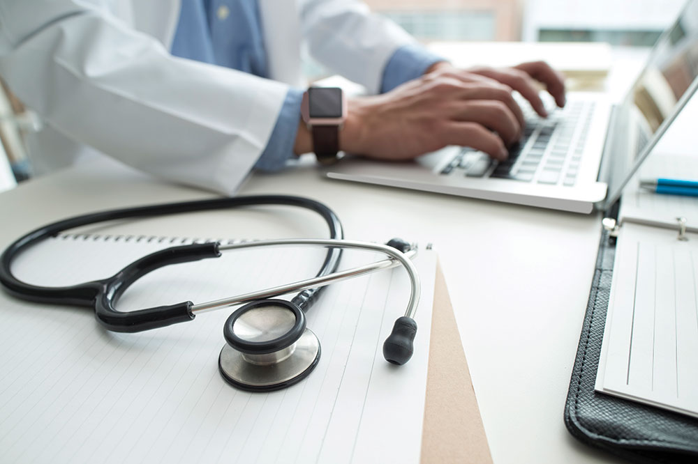 Doctors hands typing on laptop, stethoscope and notebook on the same desk