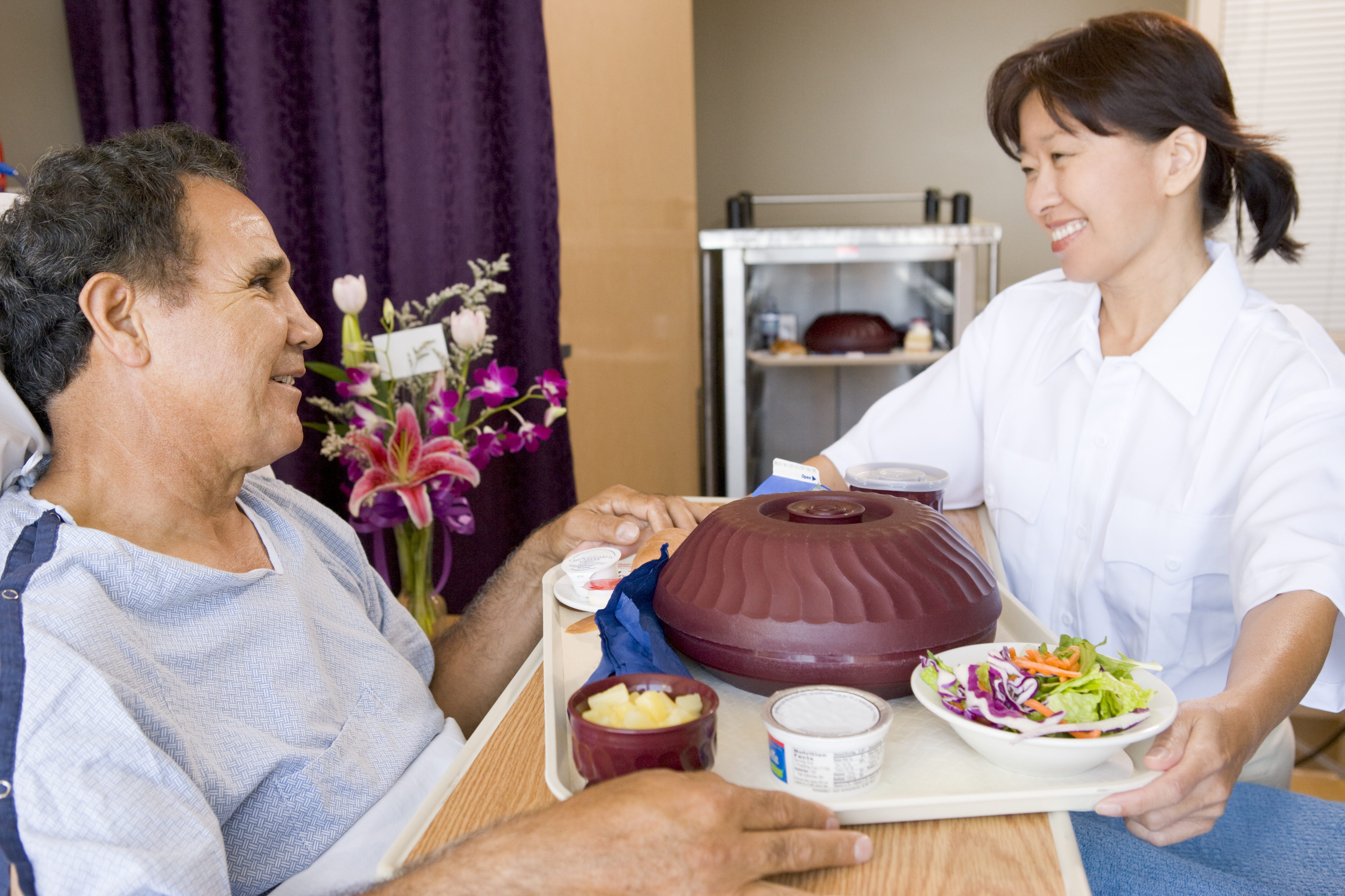 Female Asian nurse delivering a full meal tray to a hospital patient seated in bed