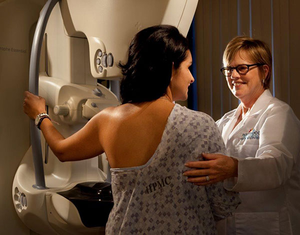 Nurse welcoming a brunette haired woman in a hospital gown over to a mammogram machine