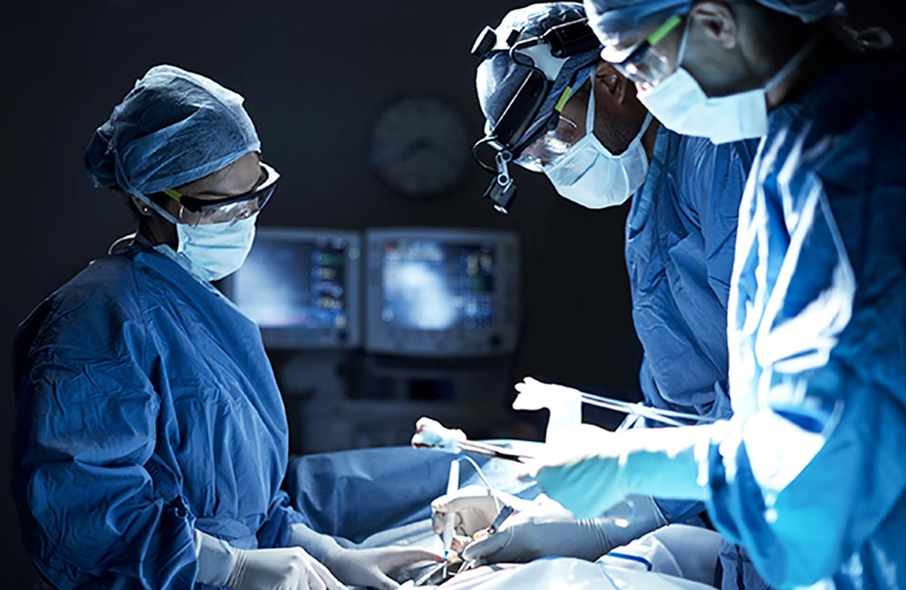 One woman adn two male surgeons with masks, gowns, and caps, leaning over a patient in the operating room