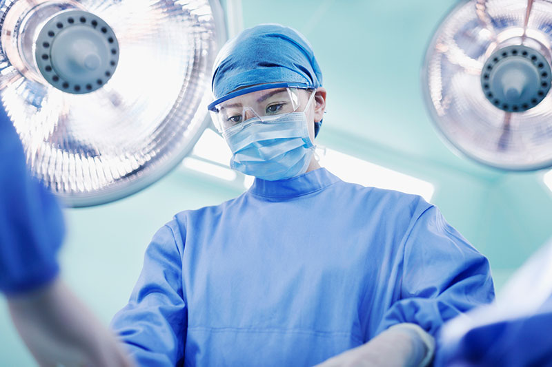 One woman adn two male surgeons with masks, gowns, and caps, leaning over a patient in the operating room