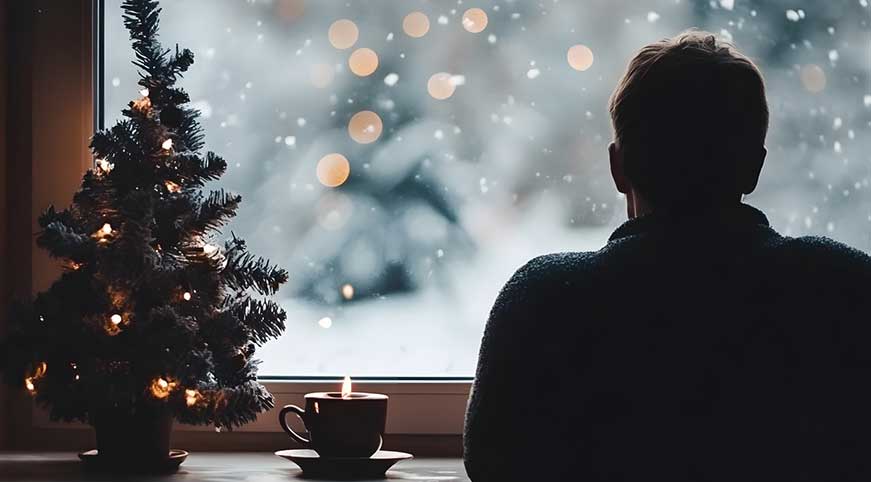 Man staring out the window at winter landscape with small Christmas tree next to him